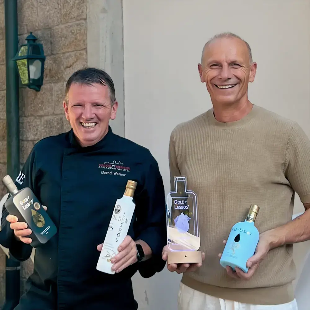 Küchenchef Bernd Werner und Produzent Anastasios Fortomaris halten mehrere Gold-of-Lesbos-Flaschen und eine Auszeichnung in den Händen